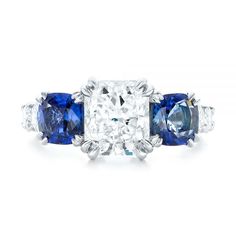 102972 Platinum Ring   2 Blue Sapphires - .91 ctw   10 Diamonds - .98 ctw   Clarity: VS2 - Color: F-G    Joseph Jewelry. This dazzling engagement ring features a radiant cut diamond in the center, with a cushion shaped blue sapphire on either side, and shared prong set diamonds down the top of the platinum Euroshank. It was created for a couple from Issaquah, WA, who already had all three stones they wanted to use for the main setting, as well as a clear vision for what they wanted the overall p Sapphire Diamond Ring With Prong Setting Cushion Cut, Sapphire Platinum Diamond Ring Cushion Cut, Sapphire Cushion Cut Diamond Ring In Platinum, Platinum Sapphire Cushion Cut Diamond Ring, Sapphire Wedding Ring With Brilliant Cushion Cut, Cushion Cut Diamond Ring With Center Stone Lab-created Sapphire, Blue Radiant Cut Diamond Ring In Platinum, Blue Diamond Ring With Round Cut, Blue Three Stone Round Cut Diamond Ring