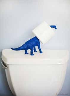 a blue toy dinosaur with a roll of toilet paper on its back sitting on top of a white toilet