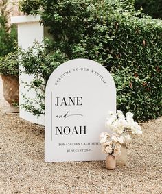 a white sign sitting on top of a gravel road next to bushes and flowers in vases