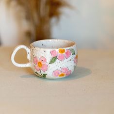 a white cup with pink flowers painted on it sitting on a table next to a potted plant