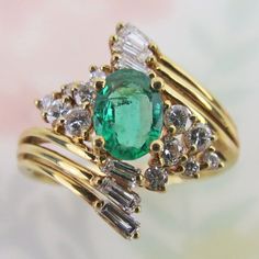 Natural diamond clusters and yellow gold swirls surround a lovely bright green emerald gem on this vintage 14k yellow gold ring. The gemstones are bright, vivid and lively. The round faceted diamonds are mounted in tiered settings. The diamond baguettes are set at the end of each yellow gold swirl, giving the ring lots of visual sparkle and movement. The ring was finished with a linear gallery and a sturdy thick gold shank band.  The oval-faceted emerald is of rich medium green color with visibl Green 14k Gold Cluster Ring, Green Cluster Diamond Ring For Anniversary, Green Emerald Cluster Ring With Diamond Accents, Green Diamond Ring Stamped 14k For May Birthstone, Yellow Gold Cocktail Ring, Emerald Gem, Gold Cocktail Ring, Gold Cocktail, Green Emerald