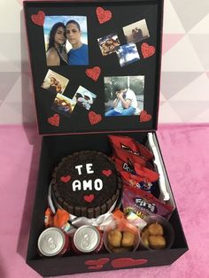 an open box with various items inside on a pink tableclothed surface, including a cake and two cans of sodas