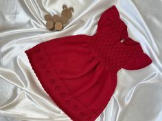 a teddy bear laying next to a red dress on a white sheet with a wooden toy