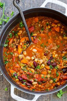 a pot filled with soup and beans on top of a wooden table next to a spoon