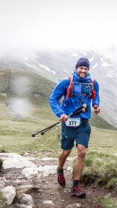 a man running up a trail in the mountains with skis on his back and poles attached to his waist