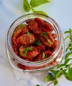 a glass jar filled with sliced tomatoes on top of a white marble counter next to green leaves