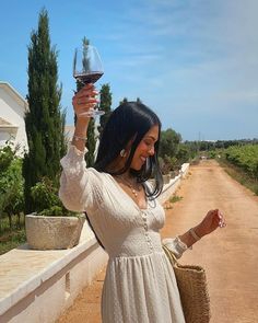 a woman in a white dress holding a wine glass up to her head while standing on a dirt road