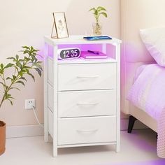 a night stand with a clock on it next to a bed and potted plant