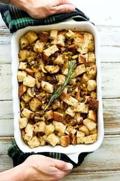 two hands holding a casserole dish filled with stuffing and vegetables on a wooden table