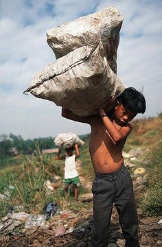 a young boy carrying a large bag over his head on the side of a dirt road