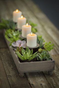 some white candles are sitting in a wooden box with succulents and greenery