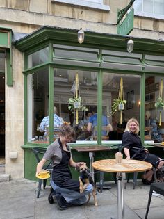 two women sitting at an outdoor table with a dog in front of the storefront