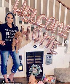 a woman holding a teddy bear standing in front of some balloons that say welcome home