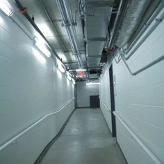 a long hallway with white walls and metal piping on the ceiling is dimly lit