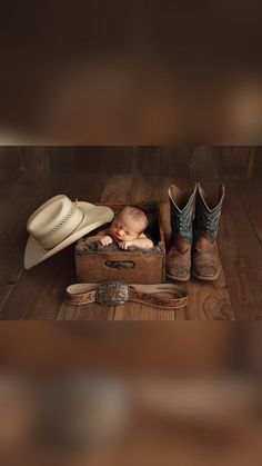 a baby is sleeping in a box with cowboy boots and other items around it on a wooden floor