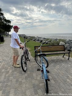 a man standing next to a bike on a brick walkway near the ocean with a bench