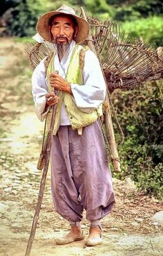 an old man with a large stick and hat walking down a dirt road in front of trees