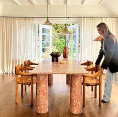 a woman standing in front of a large wooden table with chairs and vases on it