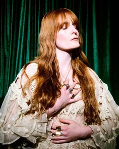 a woman with long red hair standing in front of a green curtain and holding her hands on her chest