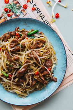 a blue plate topped with meat and noodles on top of a table next to a magazine