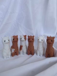 five small stuffed animals are lined up on a white cloth covered surface, one is brown and the other is white