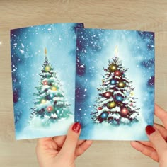 two cards with christmas trees on them in front of a wooden table and a person holding one