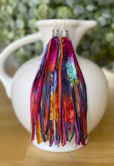 a white vase with some colorful tassels hanging from it's sides on a wooden table