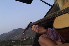 a woman playing an acoustic guitar while sitting in a vehicle with mountains in the background