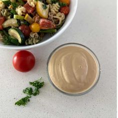 a white bowl filled with pasta salad next to a small container of dressing and a cherry tomato