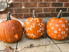 three painted pumpkins sitting on the ground next to a brick wall with white flowers