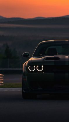 the front end of a black sports car driving on a road at sunset with mountains in the background