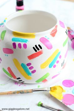 a white bowl with colorful paint splattered on it next to paints and brushes