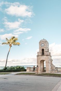 two people standing in front of a clock tower with a palm tree on the other side