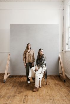 two women are sitting on chairs in an empty room with wood flooring and white walls