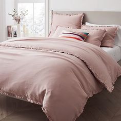 a bed with pink comforter and pillows in a white room next to a window