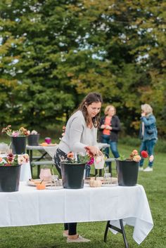 a woman is arranging flowers on a table in the grass with other people standing around