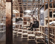 two people sitting on wooden chairs in a room filled with books and photographs, while looking at a laptop