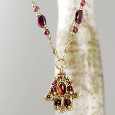 A White Metal & Brass Hamsa necklace electroplated with 24k Gold that is embellished with garnet and glass beads on a beaded chain is inspired by the Middle Eastern symbol of good luck, intended to ward off evil and protect it's wearer. This handmade piece is a one of a kind, fashionable take on a traditional cultural charm that will be perfect for any wearer. Pendant Length: .75 inches Necklace Length: Adjustable: 18 inches to 22 inches Additional Extender: 4 inches Item Number: N2734 Profe Gold Czech Glass Jewelry, Hand-strung, Hand-strung Gold Jewelry With Czech Glass, Hand-strung Gold Czech Glass Jewelry, Gold Necklaces With Round Beads For Blessing, Hand-strung Brass Jewelry For Gift, Hand-strung Brass Jewelry Gift, Handmade Garnet Beaded Necklaces, Handmade Garnet Bead Necklaces, Handmade Garnet Necklaces With Round Beads