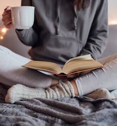 a woman is sitting on the bed and reading a book while holding a cup of coffee