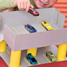 a young boy playing with toy cars in a cardboard box on the floor next to yellow poles