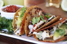 three tacos on a plate with salsa and guacamole in the background