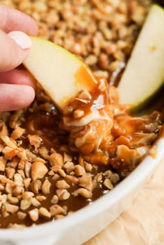 an apple being dipped with caramel sauce in a bowl filled with nuts and apples