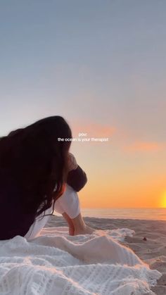 a woman sitting on top of a blanket next to the ocean at sunset with her hair blowing in the wind