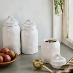 two white jars sitting on top of a counter next to apples and other food items