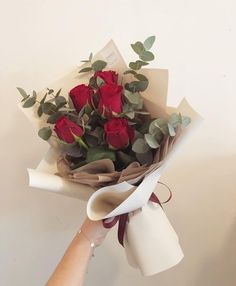 a hand holding a bouquet of red roses with greenery on the stems and wrapping paper