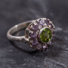 Peridot Ring set with Natural Peridot in a diamond cut & natural green color, at size 7mm together with Natural Amethyst at size 3mm each (2.3 Cts combined) from Brazil.Solid 925 Sterling Silver ☞ made to last.Click here for ☞ Matching PendantClick here for ☞ Matching EarringsClick here for ☞ Flower Collection Details:• Natural Peridot & Amethyst sourced from Brazil• Peridot: 7mm, 1.3 Cts, diamond cut• Amethyst: 3mm each, 1 Ct total, diamond cut• Dimensions: Band width ≈ 2.3mm, thickness ≈ 1.3mm Round Peridot Gemstones Fine Jewelry, Green Multi-stone Cluster Promise Ring, Green Sterling Silver Amethyst Ring For Wedding, Green Amethyst Rings In Fine Jewelry Style, Green Amethyst Promise Ring In Fine Jewelry Style, Green Multi-stone Cluster Ring For Promise, Green Peridot Ring With Center Stone, Green Amethyst Ring With Accent Stones For Promise, Green Amethyst Ring In Sterling Silver