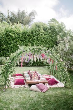 a picnic table with pink pillows and flowers on it in the middle of a lush green yard