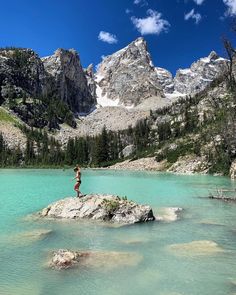 a person standing on top of a rock in the middle of a lake surrounded by mountains