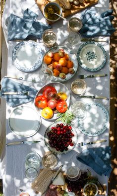 a table with plates, bowls and fruit on it in the shade under a tree