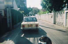 an old car driving down the street in front of a fenced off area with trees